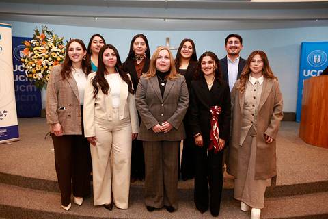 Un grupo de mujeres y un hombre posando en un evento oficial con una decoración de flores al fondo.