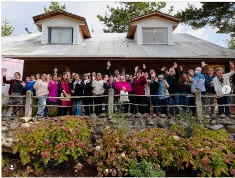 Un grupo grande de personas posando y alegremente saludando frente a una casa de campo en un día soleado.
