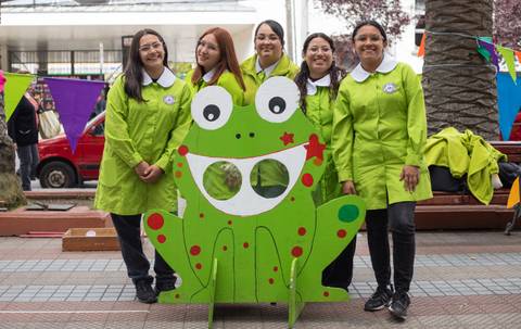 Un grupo de cinco mujeres posando alegres frente a un fondo colorido con una figura de rana grande.