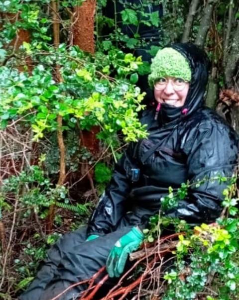 Una persona sentada entre arbustos verdes, con un gorro de lana y vestido de negro, disfrutando de la naturaleza.