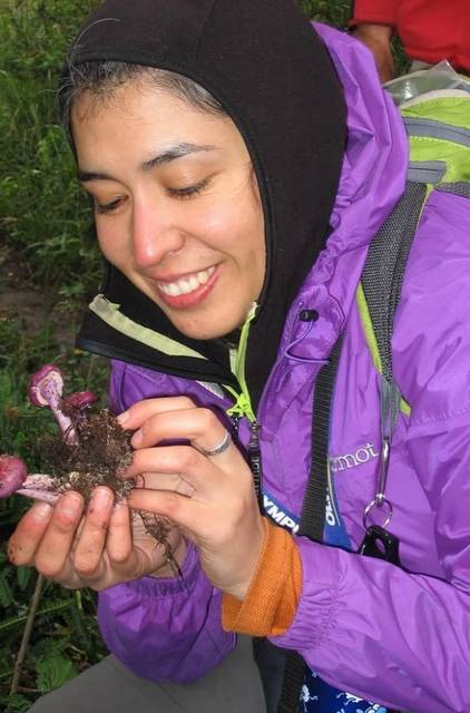 Una mujer sonriente sostiene con cuidado un hongo morado en medio de la naturaleza.