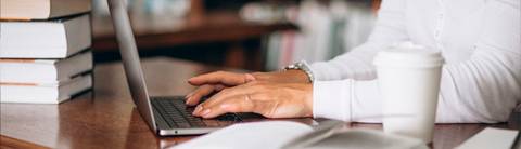 Una persona está escribiendo en un portátil sobre una mesa con libros y una taza de café.