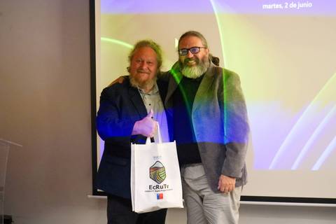 Dos hombres posan juntos sonrientes en un evento, uno sosteniendo una bolsa con el logo de ECRUTv.