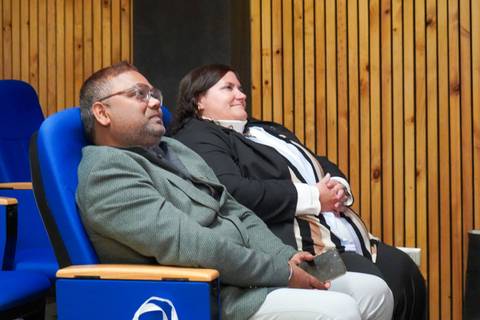 Un par de personas sentadas en asientos azules, observando una presentación.