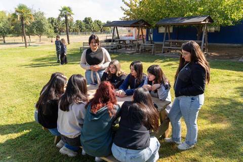 Un grupo de estudiantes se reúne alrededor de una mesa en un parque, disfrutando de una actividad al aire libre.