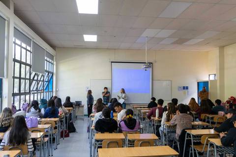 Una clase con estudiantes atentos mientras tres mujeres presentan un tema en la pizarra.