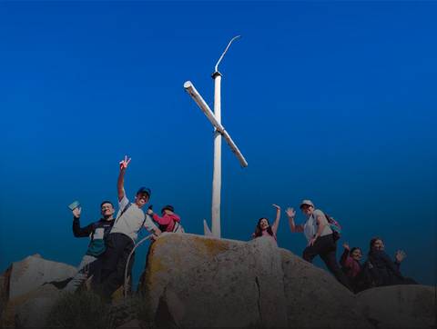 Un grupo de amigos celebrando en la cima de una roca junto a una cruz blanca bajo un cielo despejado.