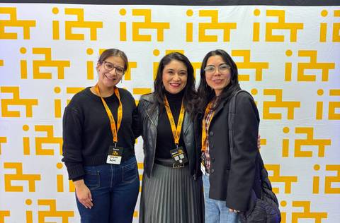 Tres mujeres sonrientes posando juntas en un evento con un telón de fondo amarillo y blanco.