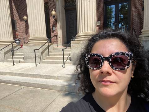 Una mujer con anteojos oscuros sonríe frente a un edificio con columnas y escaleras en un día soleado.