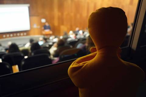 Un muñeco amarillo observa una presentación desde la ventana en una sala llena de gente.