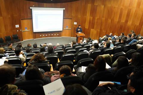 Una charla llena de gente en un auditorio, con una presentación en pantalla.