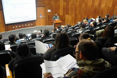 Un expositor presenta en un auditorio mientras la audiencia toma apuntes.