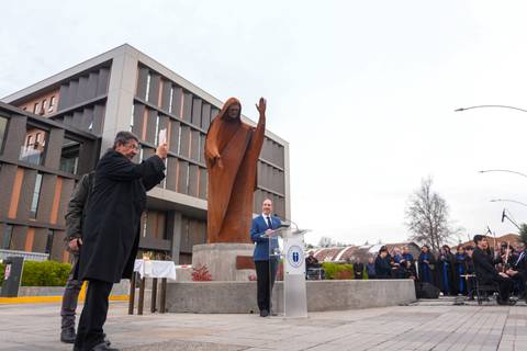 En la imagen se ve a un político dando un discurso frente a una escultura mientras un hombre la fotografía.