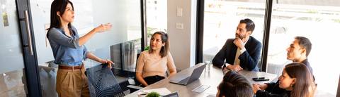 Una mujer está presentando en una reunión con varias personas escuchando atentamente.