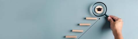 Una mano sostiene una lupa enfocando un símbolo de graduación sobre unas escaleras de madera que representan el progreso académico.