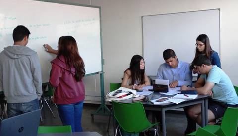Un grupo de estudiantes trabaja en un aula, algunos escriben en una pizarra y otros colaboran en una mesa.