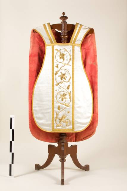 Una vestidura ceremonial con bordados en oro y detalles florales, colocada sobre un soporte de madera.