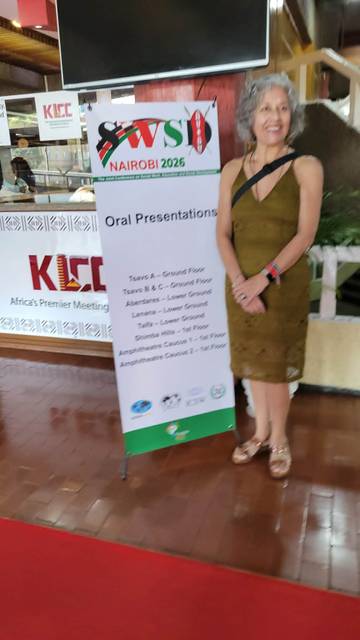 Una mujer sonriente posa junto a un cartel de presentaciones orales en un evento.