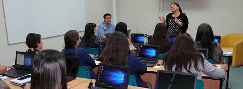 Una profesora está dando una charla mientras sus alumnos escuchan atentamente en un salón de clases.