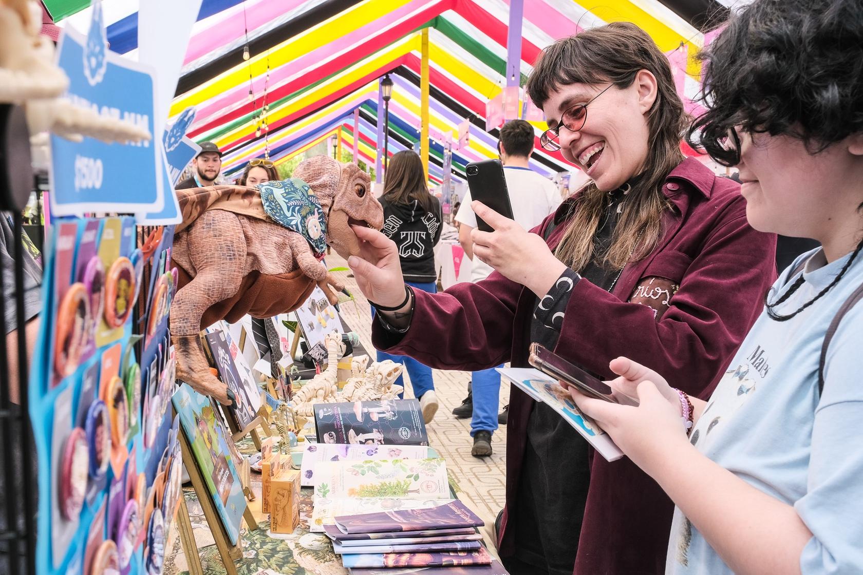 En una feria llena de coloridos stands, dos amigas se ríen mientras observan una figura de dinosaurio.