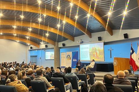 Un evento con una gran cantidad de personas sentadas, escuchando a un expositor en un auditorio iluminado.