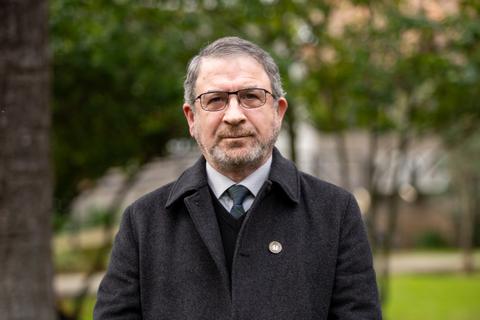 Un hombre mayor de cabello gris, usando gafas y un abrigo oscuro, posa frente a un fondo natural.