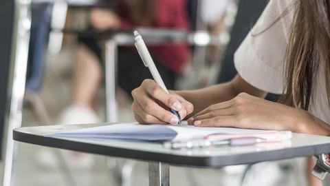 Una persona está escribiendo con un bolígrafo sobre una hoja de papel en un entorno de aula.