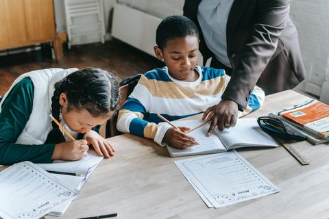 Dos niños están estudiando en una mesa mientras un adulto les ayuda.