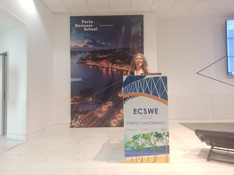 Una mujer sonriente se encuentra detrás de un podio en un salón de conferencias con un fondo de la ciudad de Oporto.