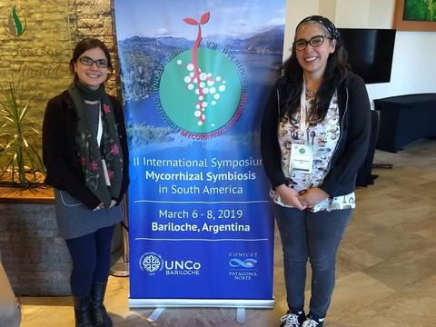 Dos mujeres posan sonrientes junto a un cartel del Simposio Internacional sobre Simbiosis Micorrízica en Bariloche.