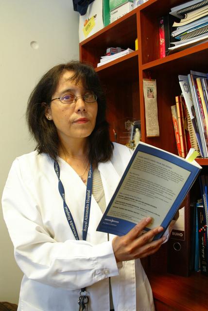 Una mujer con bata de laboratorio sostiene un libro frente a una estantería llena de documentos.