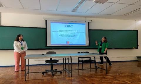 Una presentación en una sala con dos mujeres de pie junto a una mesa frente a una pantalla con contenido relacionado a la UCM.
