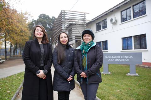 Tres mujeres posan frente a un edificio académico rodeado de árboles en un día nublado.