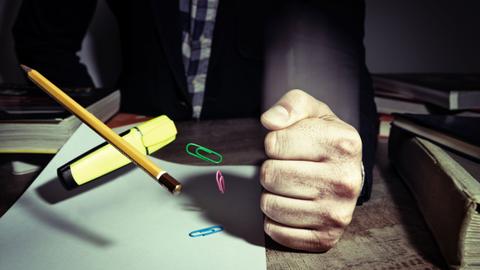 Una mano cerrada sostiene un lápiz mientras varios sujetapapeles flotan sobre una hoja de papel en un ambiente oscuro.