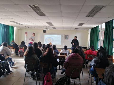 Una clase en un aula con un profesor hablando frente a una pantalla y varios estudiantes escuchando.