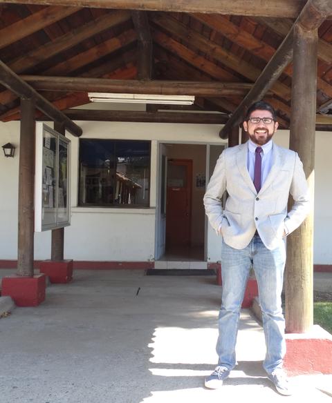 Un hombre sonriente en un traje claro se encuentra de pie frente a una estructura con techo de tejas.