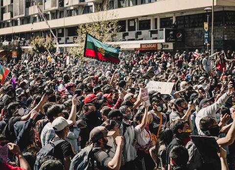 Una multitud de personas protesta en una plaza, ondeando banderas y mostrando carteles.