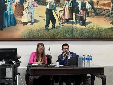 Una presentación en mesa con dos ponentes frente a un mural que representa escenas históricas.