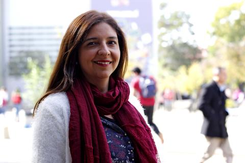 Una mujer sonriente, vestida con un suéter blanco y una bufanda roja, se encuentra de pie en un entorno urbano con gente al fondo.