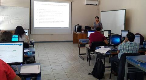 Un aula con estudiantes asistiendo a una clase impartida por un docente frente a una pantalla con contenido proyectado.