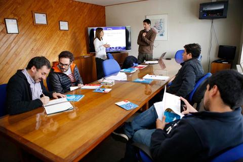 Un grupo de personas se encuentra reunido en una sala de trabajo discutiendo y revisando materiales en una mesa.