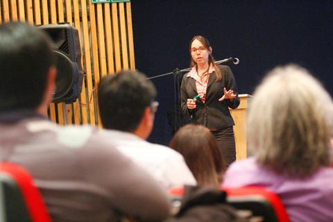 Una mujer está presentando en un evento mientras un grupo de personas la observa atentamente.