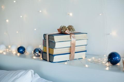 Una pila de libros decorados con un lazo y adornos navideños sobre una repisa iluminada con luces.