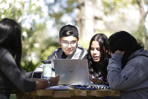 Un grupo de cuatro jóvenes se encuentra reunido al aire libre, trabajando juntos en una computadora portátil.