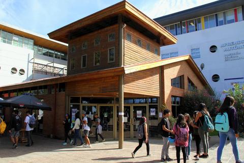 La imagen muestra un edificio de madera con personas caminando a su alrededor en un ambiente escolar.
