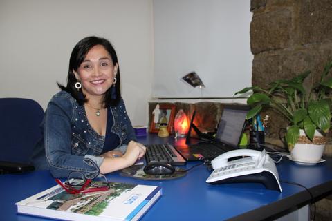 Mujer sonriente sentada en un escritorio con un ordenador y una planta.