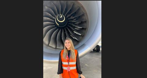 Una mujer sonriente con un chaleco reflectante naranja está de pie frente a un motor de avión grande.