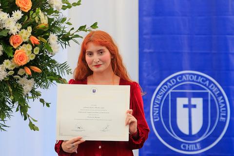 Una persona sostiene un certificado frente a un fondo decorado con flores y un logo de la universidad.