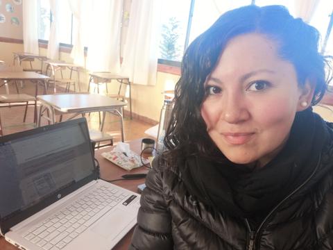 Una persona sonriente se toma una selfie frente a una computadora portátil en un aula.