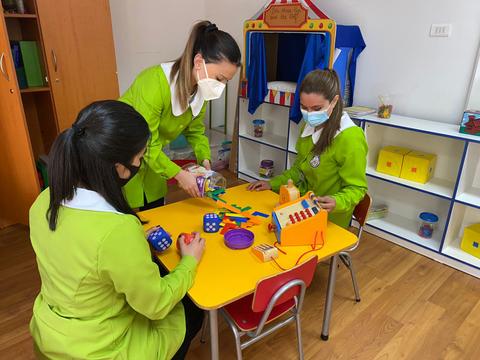 Tres personas en un aula infantil están jugando y aprendiendo con juguetes educativos en una mesa.